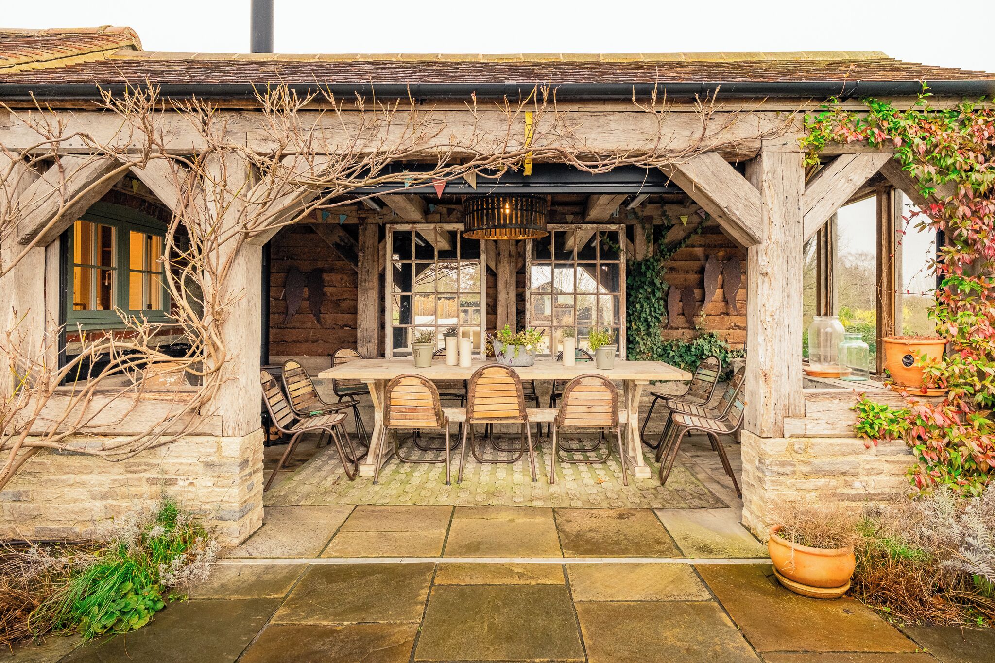 A rustic outdoor dining area with a stone table and wooden chairs, surrounded by stone walls and covered with a wooden pergola.