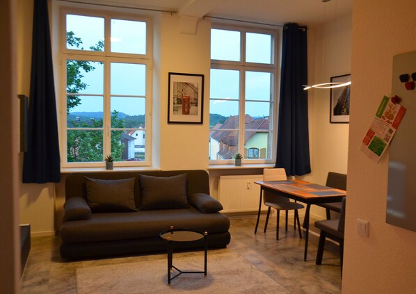 A living room with a sofa, a small round table, a dining table with chairs, large windows with curtains, and a framed picture on the wall.