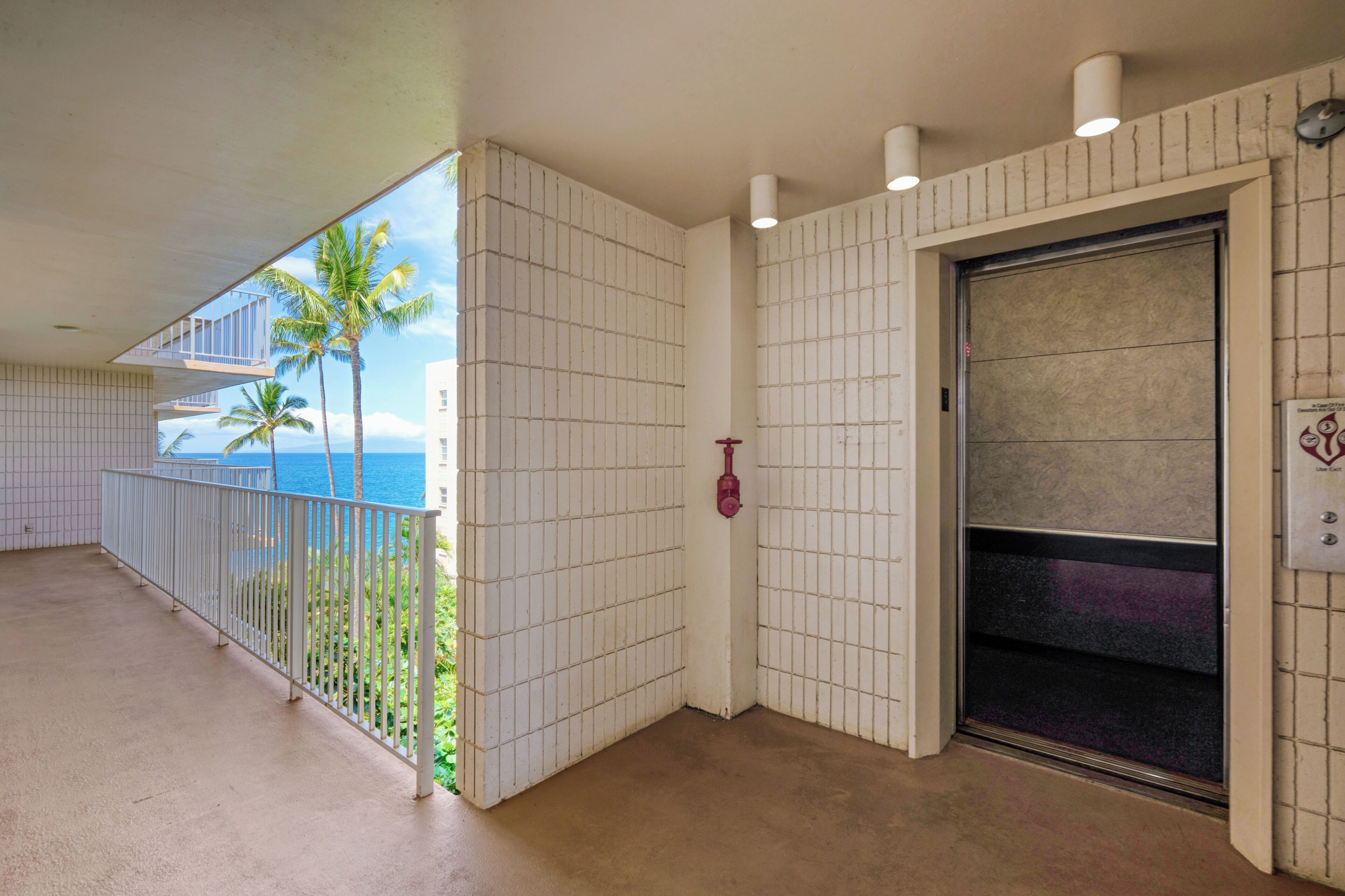 A hotel corridor with a view of the ocean and palm trees, a white tiled wall, and a closed elevator.