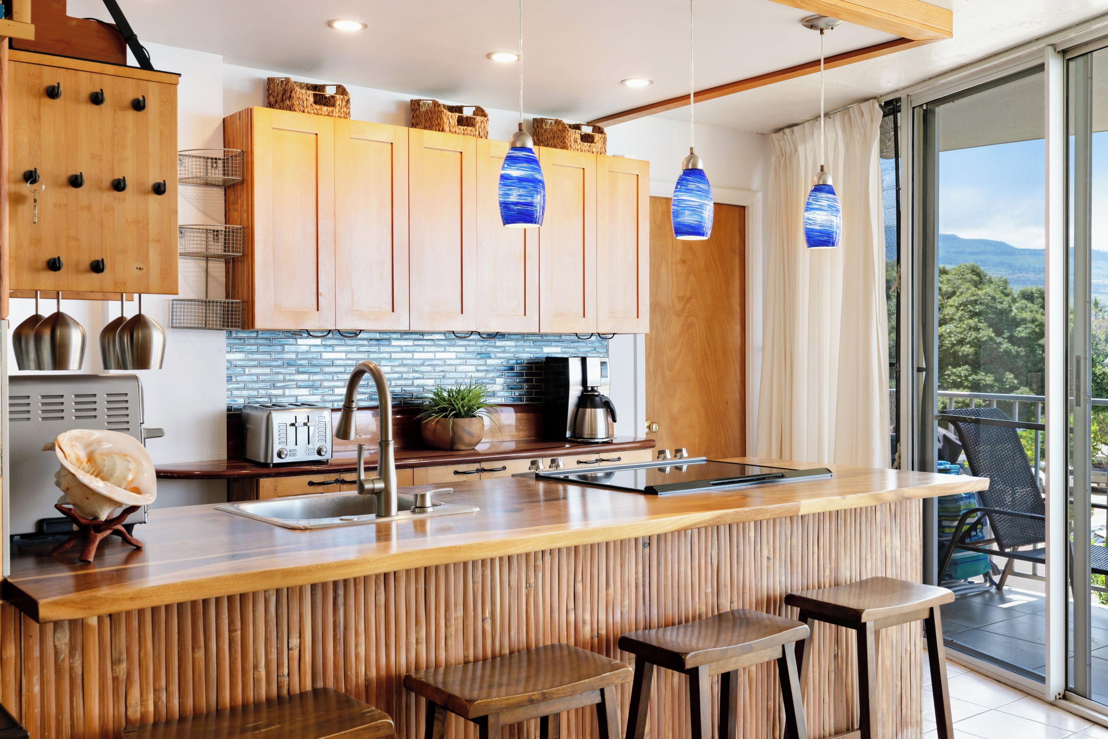 A modern kitchen with wooden cabinets, a central island with stools, a built-in toaster, and a view of the outdoors through large windows.