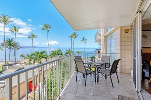 A balcony with a table and chairs, overlooking a beach with palm trees and the ocean. - Kamaole Nalu 401 - Oceanfront Complex Sunset Views (Kihei)