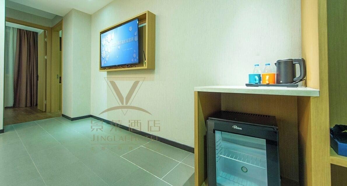 Camera d'albergo con televisore a parete, mini-frigorifero e un ripiano con bollitore e bottiglie.