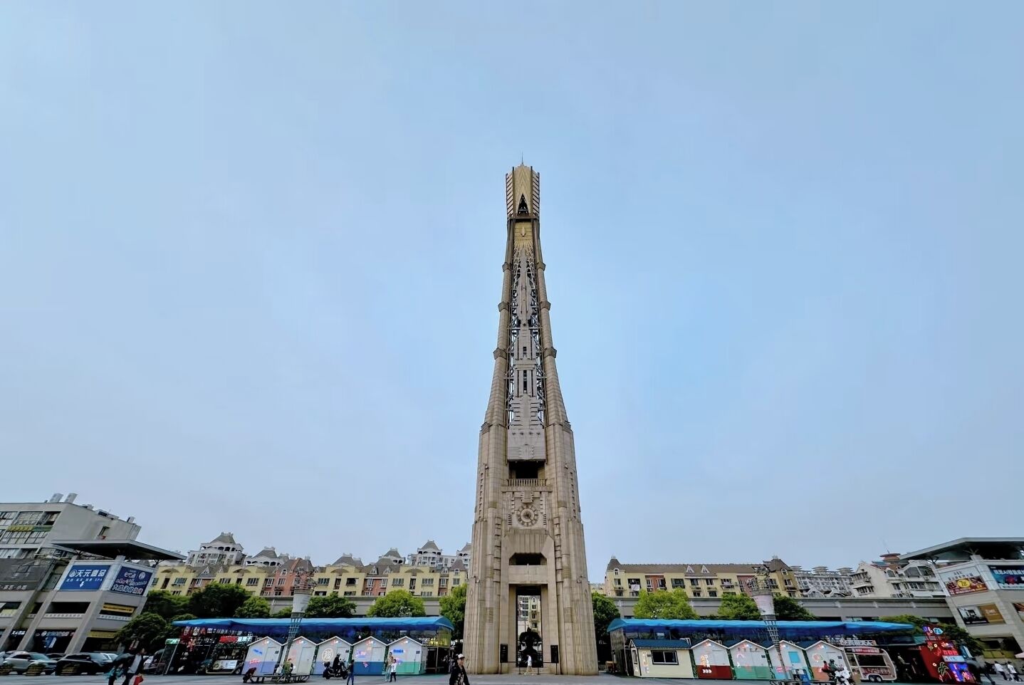 Una torre alta y esbelta con un reloj en la parte superior, rodeada de edificios y un refugio cubierto para autobuses.