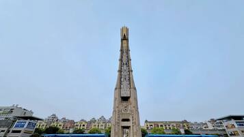 Una torre alta y esbelta con un reloj en la parte superior, rodeada de edificios y un refugio cubierto para autobuses.