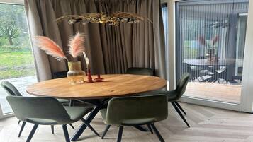 A modern dining area with a round wooden table, four green chairs, a decorative vase with pampas grass, and a unique chandelier.