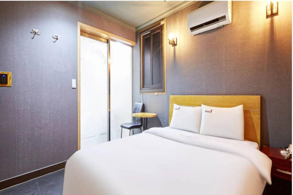 A hotel room with a large bed, a small table, a chair, and an air conditioning unit.