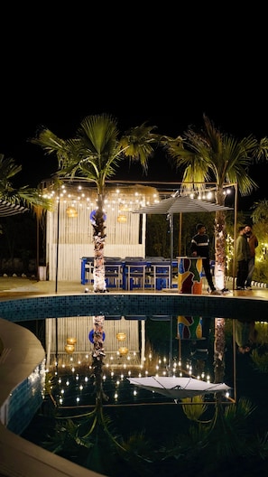 Un área junto a la piscina con piscina, palmeras y un espacio de descanso con sillas azules.