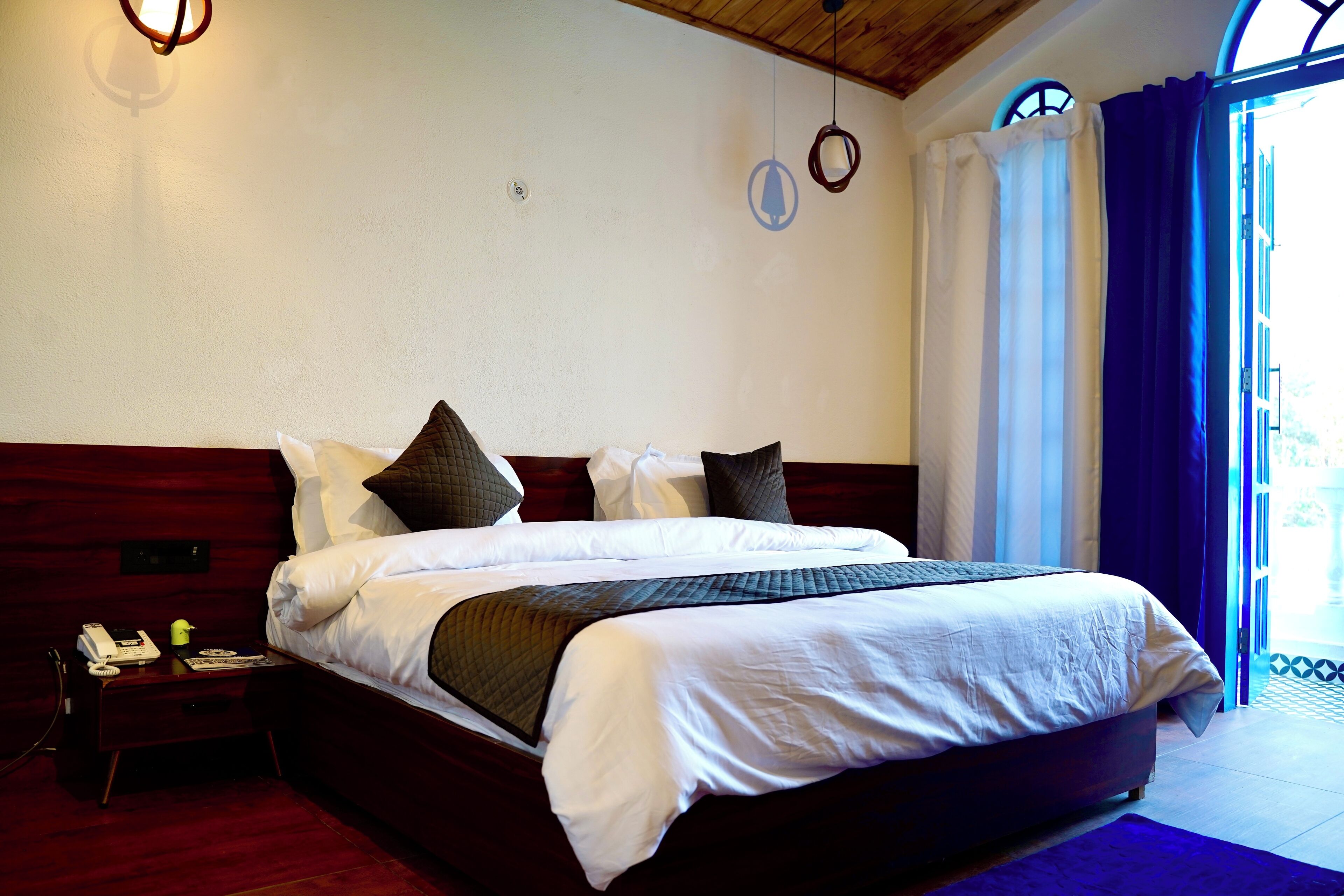 Una cama bien hecha con sábanas blancas y un cabecero oscuro, una mesita de noche con una lámpara, y vistas al exterior a través de una ventana con cortinas.