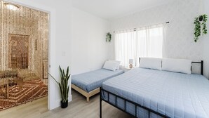 Ein Schlafzimmer mit zwei Betten, einem Fenster mit durchscheinenden Vorhängen, einer hängenden Pflanze und einem gewobenen Raumteiler.