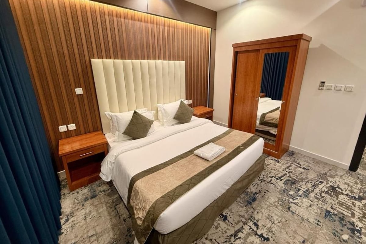 Quarto de hotel com uma cama grande, cabeceira de madeira, mesa de cabeceira, guarda-roupa e um tapete estampado.