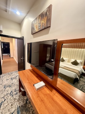 A hotel room with a large mirror, a flat-screen TV, a wooden desk, and a bed with white bedding and green pillows.
