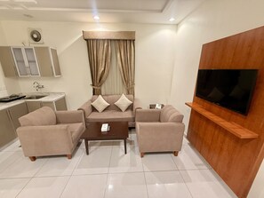 A modern living room with a sofa set, a coffee table, a TV mounted on the wall, and a kitchen area with cabinets and a sink. - Milestone Rise (Jeddah)