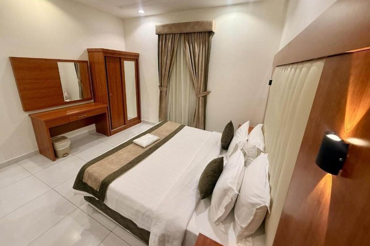 Quarto de hotel com cama, guarda-roupa, espelho, janela com cortinas e luminária fixada na parede.