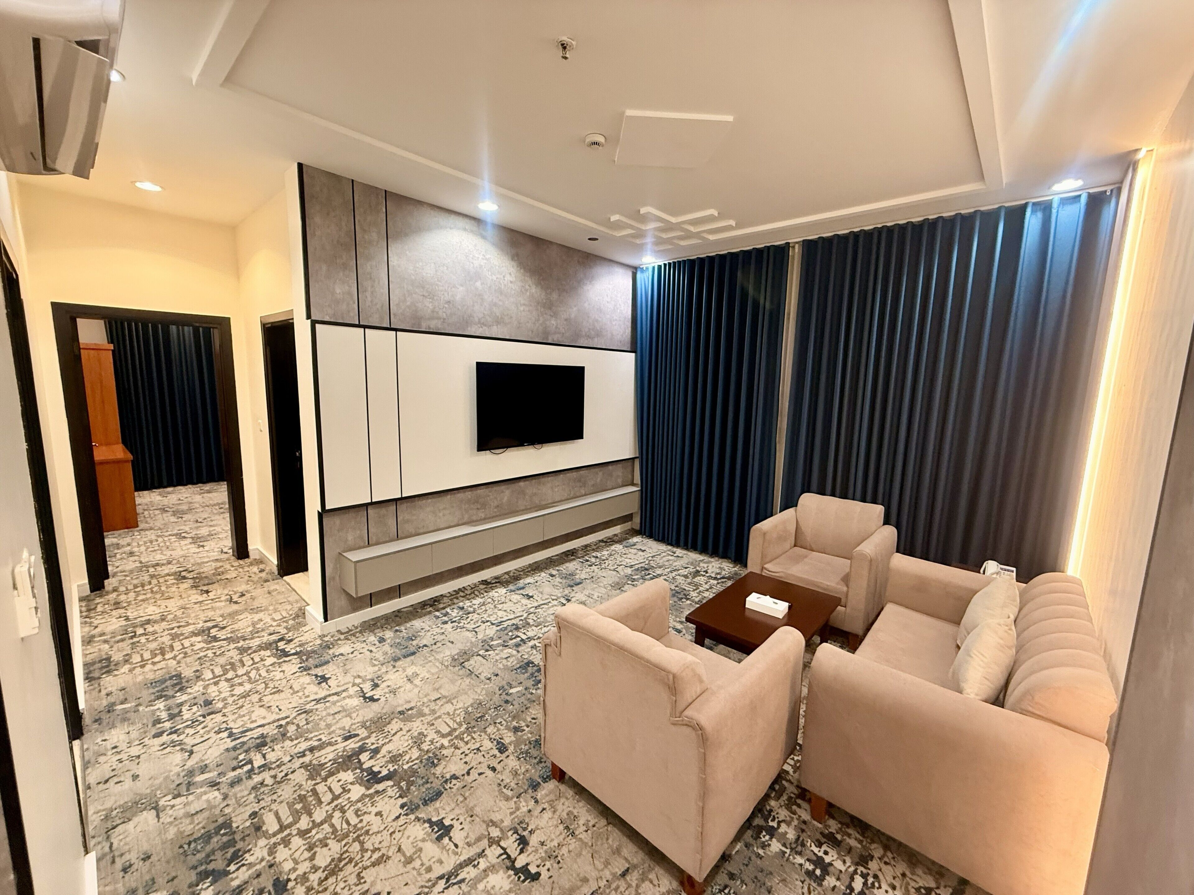 A modern living room with a flat-screen TV mounted on a wall, a patterned carpet, a beige sofa, and a coffee table.