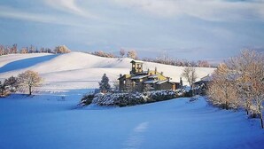 A snow-covered landscape with a building featuring a tower and multiple windows, surrounded by snow-covered trees and hills. - Il Castelletto di Gomo  (Godiasco)