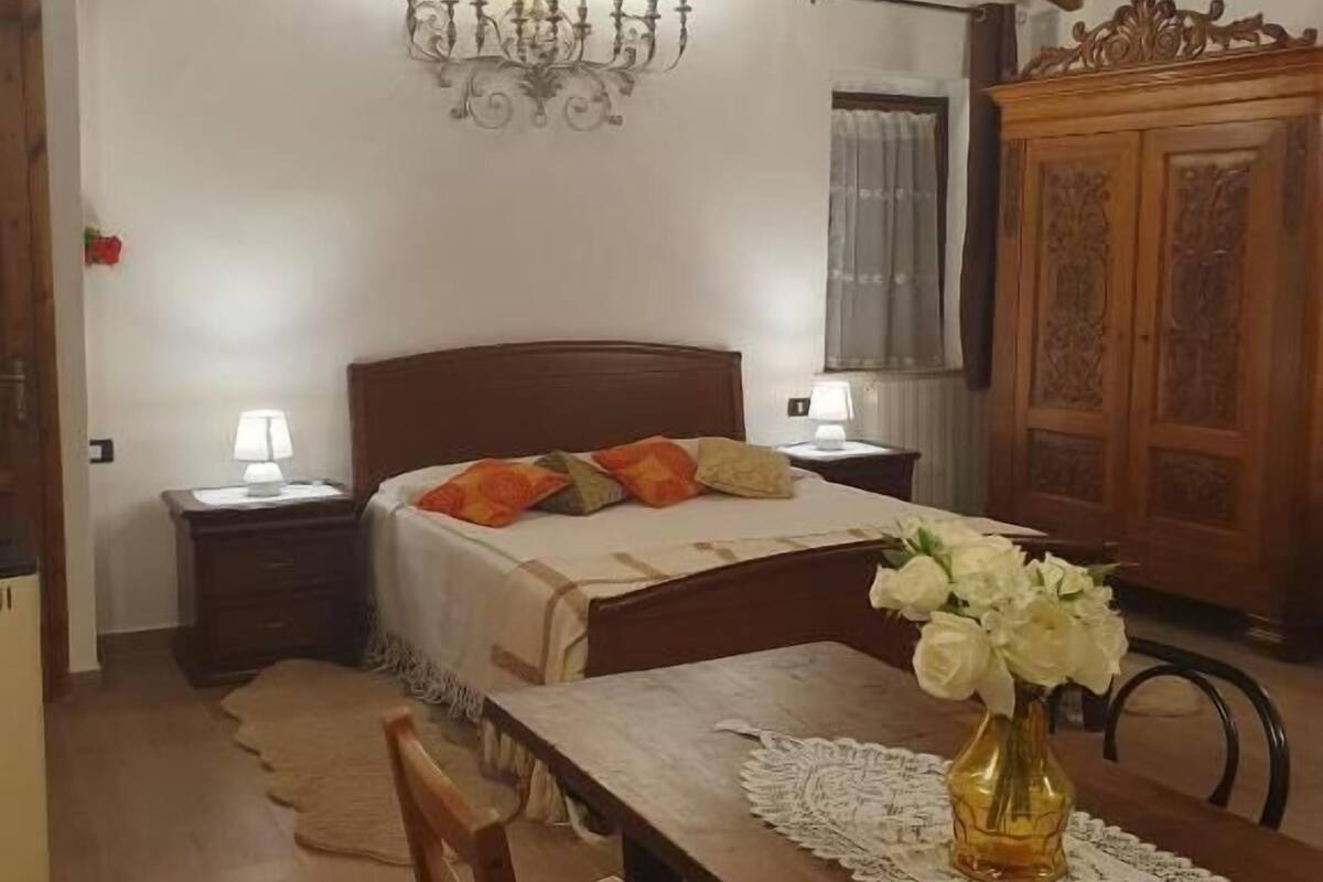 Un dormitorio con cama de madera, una mesita de noche con lámpara, una araña, un armario de madera y una mesa de comedor con sillas.