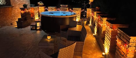 A hot tub surrounded by wicker furniture and stone walls, with string lights creating a warm ambiance.