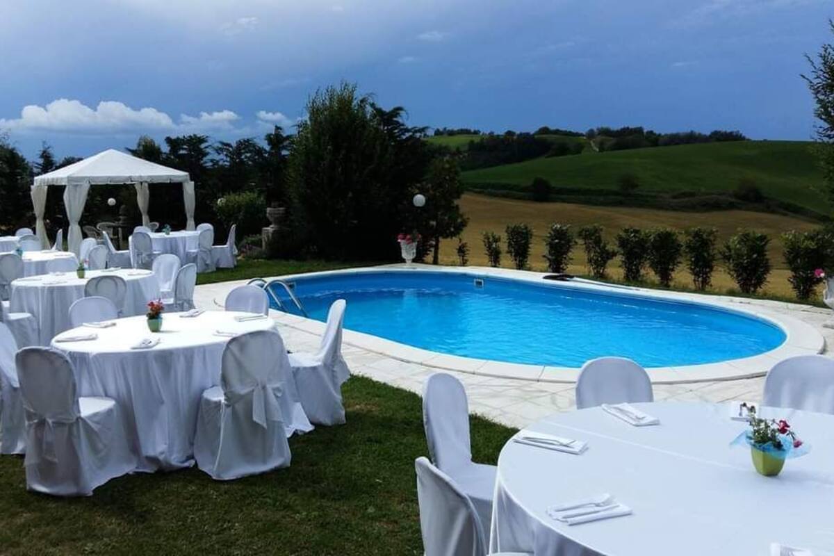 Zona exterior junto a la piscina con mesas y sillas redondas, un área de descanso cubierta y vistas al campo.