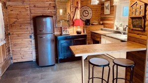 A rustic kitchen with wooden walls, a stainless steel refrigerator, a wooden table with stools, and a clock on the wall.