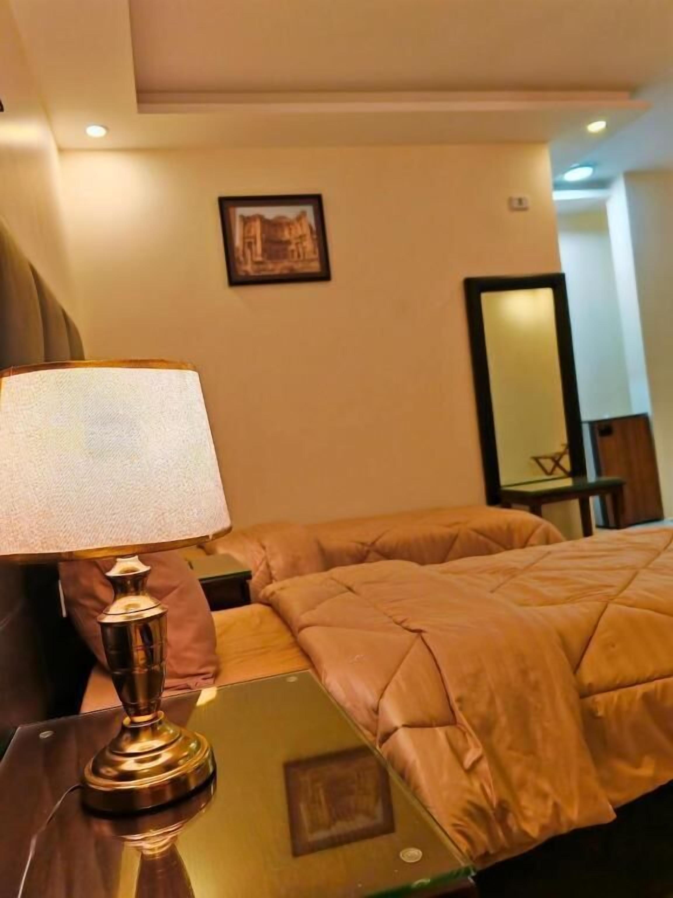 A hotel room with a bed, a desk with a lamp, a laptop, a framed picture on the wall, and a mirror.
