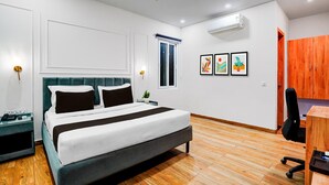 A bedroom with a bed, bedside tables, a desk, a chair, a ceiling fan, an air conditioning unit, a window, and artwork on the wall. - Townhouse Oak Sector 18 Metro Station (Noida)
