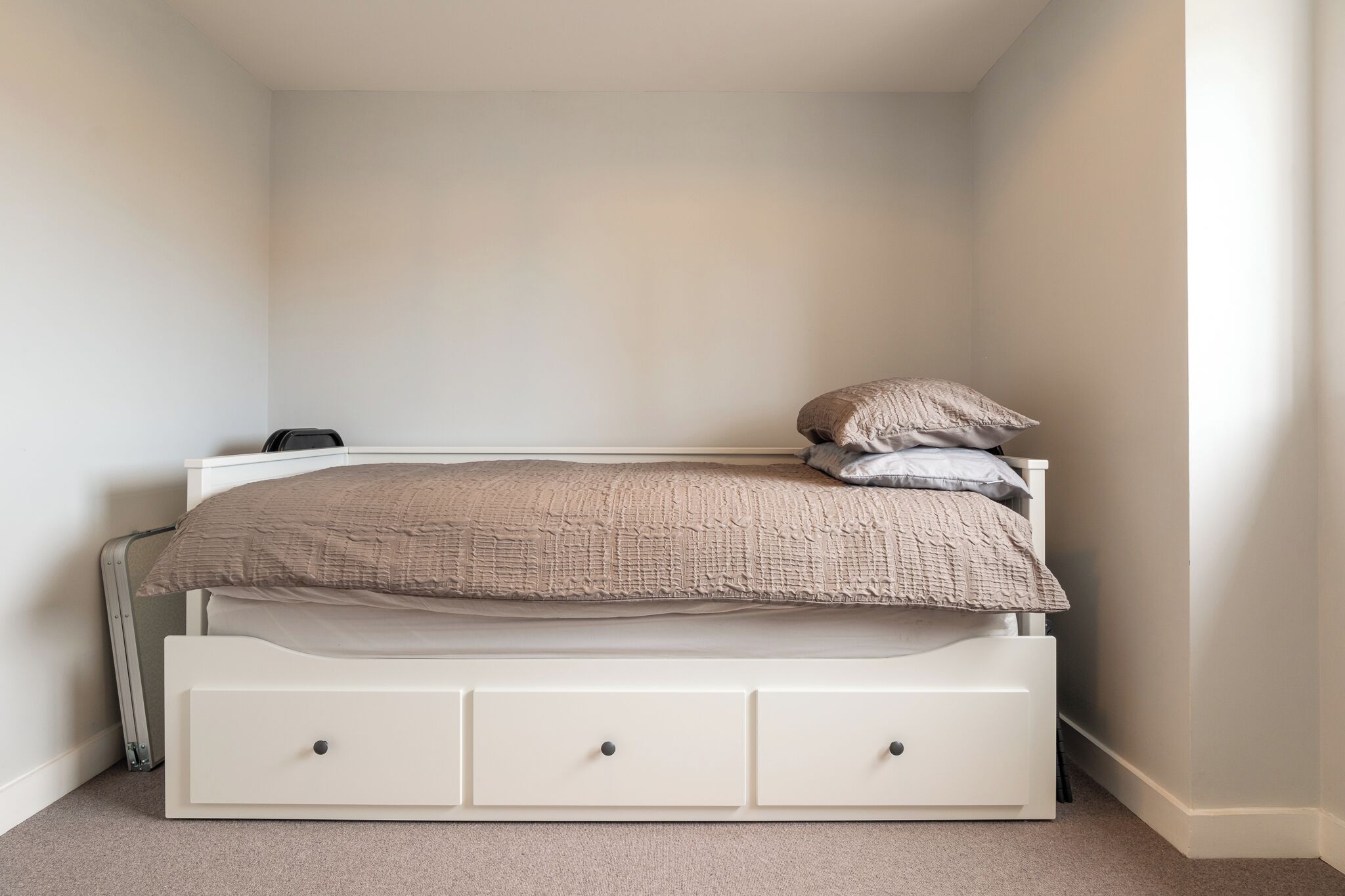 Un lit simple avec un cadre blanc et trois tiroirs, une couverture beige et deux oreillers.