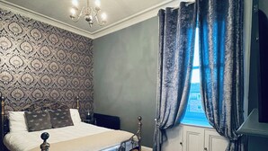 Une chambre à coucher avec un lit, un lustre, un papier peint à motifs, une fenêtre avec des rideaux et un plancher en bois.