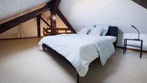 Un dormitorio con techo inclinado, una cama con colcha blanca, una silla y un escritorio con lámpara.