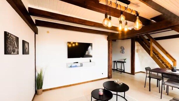 Una sala de estar moderna con televisión de pantalla plana, vigas de madera, un comedor con mesa y sillas, y una escalera.