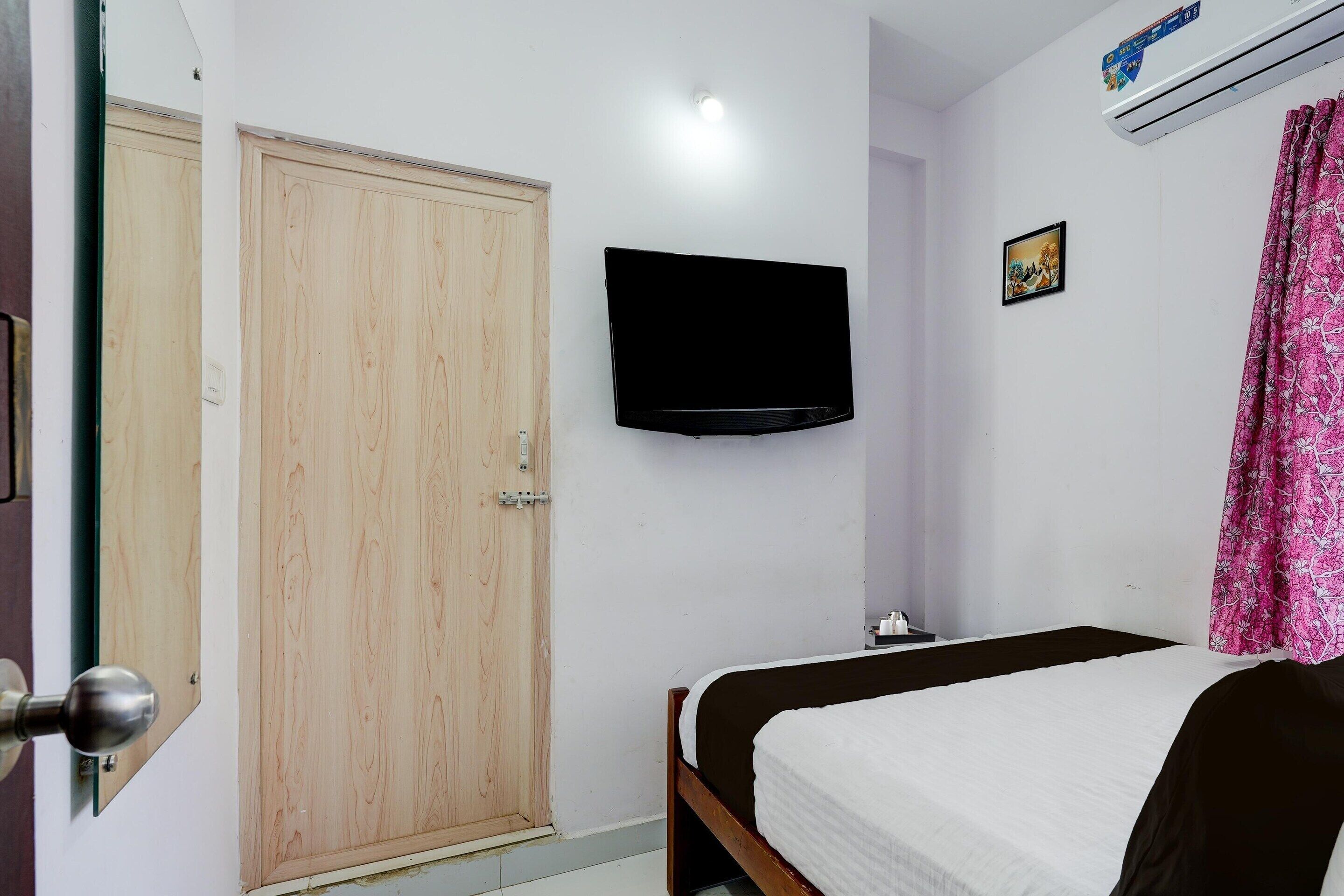 Een slaapkamer met een houten deur, een flatscreen-televisie aan de muur, een bed met een wit en zwart dekbedovertrek en een roze bloemen gordijn.