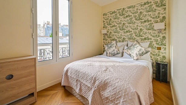 Un dormitorio con cama, mesita de noche, lámpara y ventana con vistas a edificios.