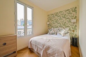 Ein Schlafzimmer mit einem Bett, einem Nachttisch, einer Lampe und einem Fenster mit Blick auf Gebäude.