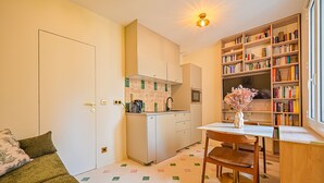 Un espacio habitable compacto con kitchenette, una zona de comedor con mesa y sillas, una estantería y un sofá con cojines.