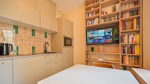 Una cocina moderna con encimera blanca, microondas y cafetera. Hay una estantería llena de libros y un televisor mostrando una videoconferencia.