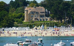 A beach with a stone building in the background, boats in the water, and beach umbrellas. - Port Manech: Charming house 100 meters from the beach, sleeps 10, exceptional garden (Névez)