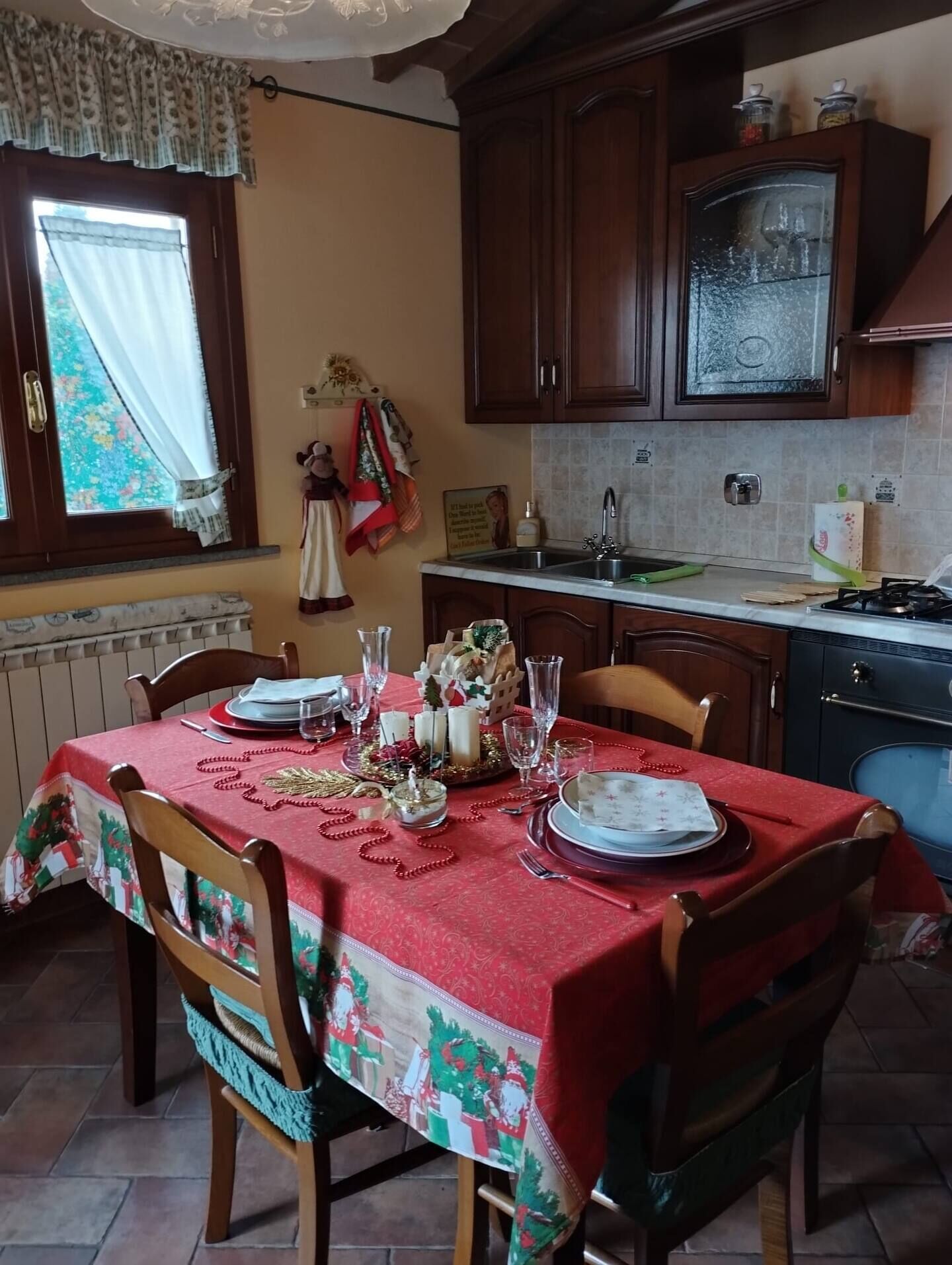 A dining area with a table set for four, featuring a red tablecloth with a Christmas theme. There are wooden chairs, a window with curtains, and a kitchen area with cabinets and a sink.