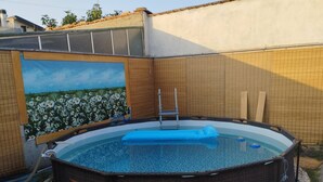 Une piscine ronde hors sol, dotée d’un escalier, entourée d’une bordure de galets et ornée d’une fresque représentant un champ de fleurs sur le mur adjacent.