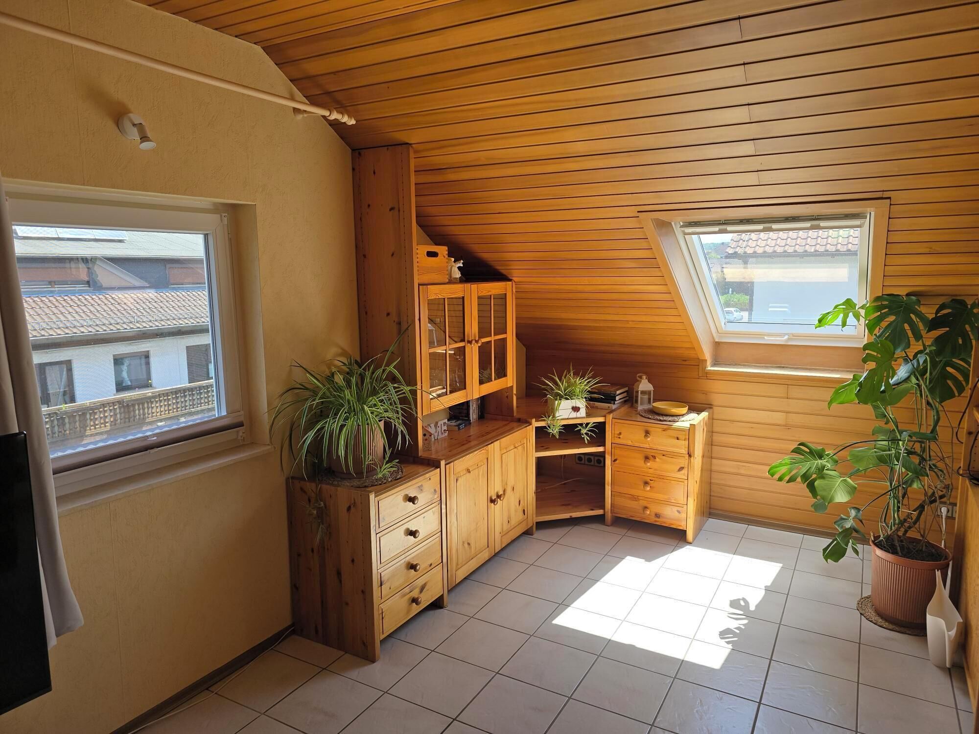 Et lille, hyggeligt rum med trævægge og skråt loft, med et vindue, et træskab og flere krukkeplanter.