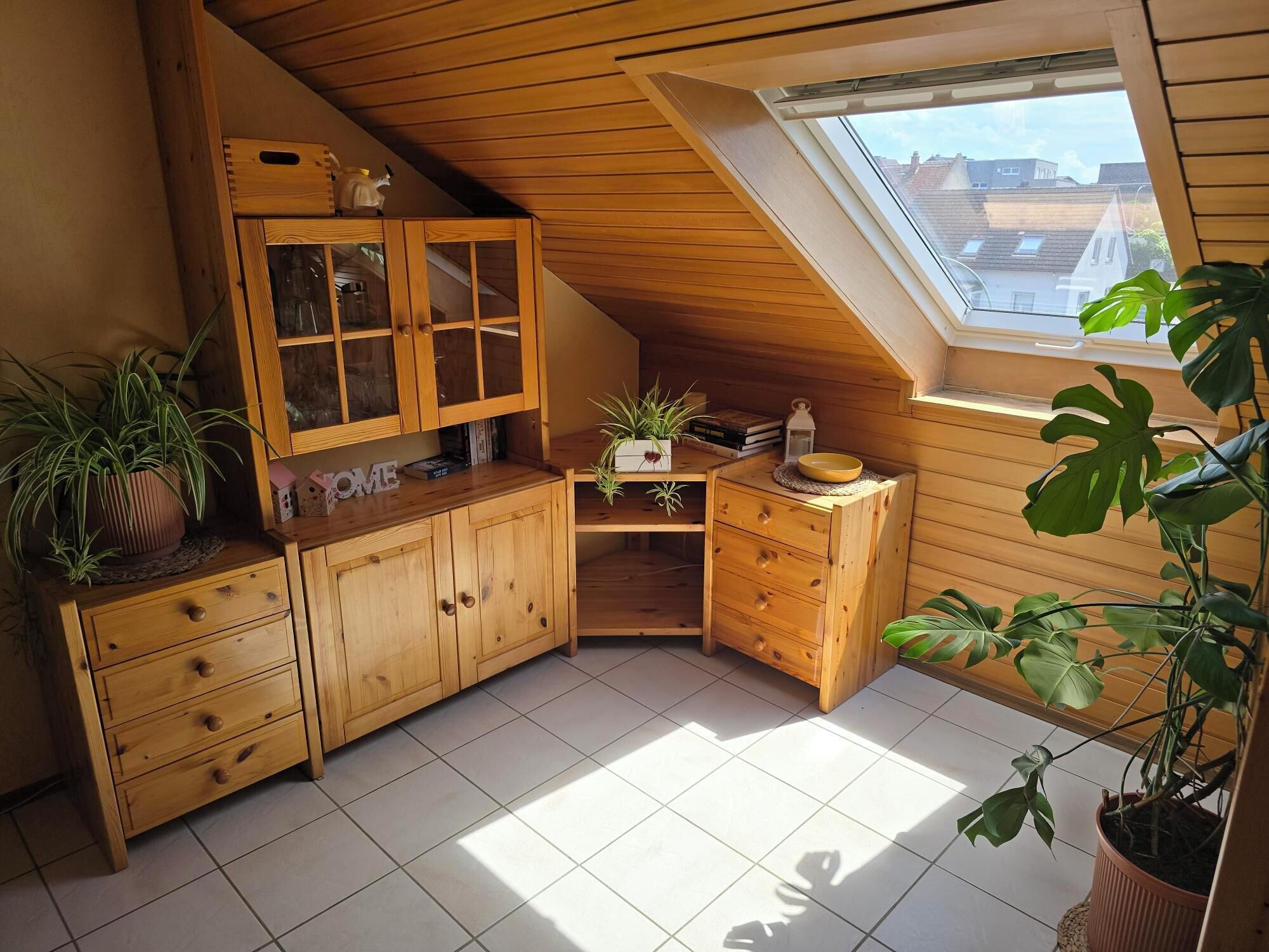 Een knus zolderkamertje met houten kasten, een dakraam en diverse planten.