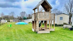 Een houten speelhuis met pannendak, een glijbaan en een schommel, gelegen in een grasveld met een bankje en een klein gebouw op de achtergrond.