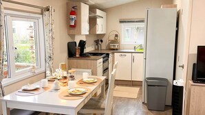 Een kleine keuken met een kookplaat, spoelbak en koelkast. Er staat een eettafel gedekt met borden en een vaas met bloemen.