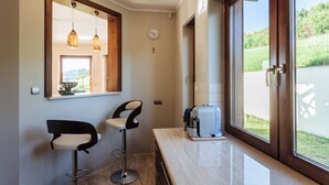 Eine moderne Küche mit zwei Barhocker, einer Kaffeemaschine und einem großen Fenster mit Blick auf Grünflächen.