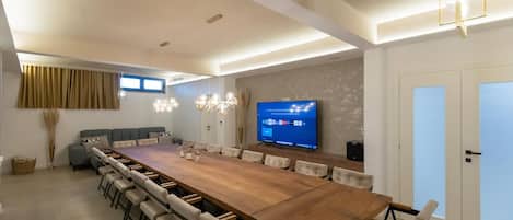 A modern living room with a large wooden dining table, comfortable seating, a flat-screen TV, and ambient lighting.