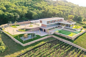 Uma casa moderna de dois andares com telhado plano, cercada por um muro de pedra e um jardim.