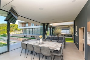 Uma área de jantar moderna com uma mesa grande e cadeiras, janelas amplas e vista para um edifício e vegetação lá fora.