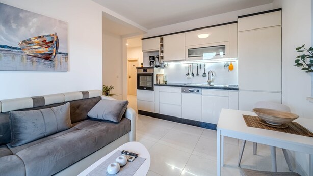 Una sala de estar moderna con un sofá gris, una mesa de centro y una mesa de comedor. Se ve una cocina con armarios blancos y electrodomésticos modernos al fondo.