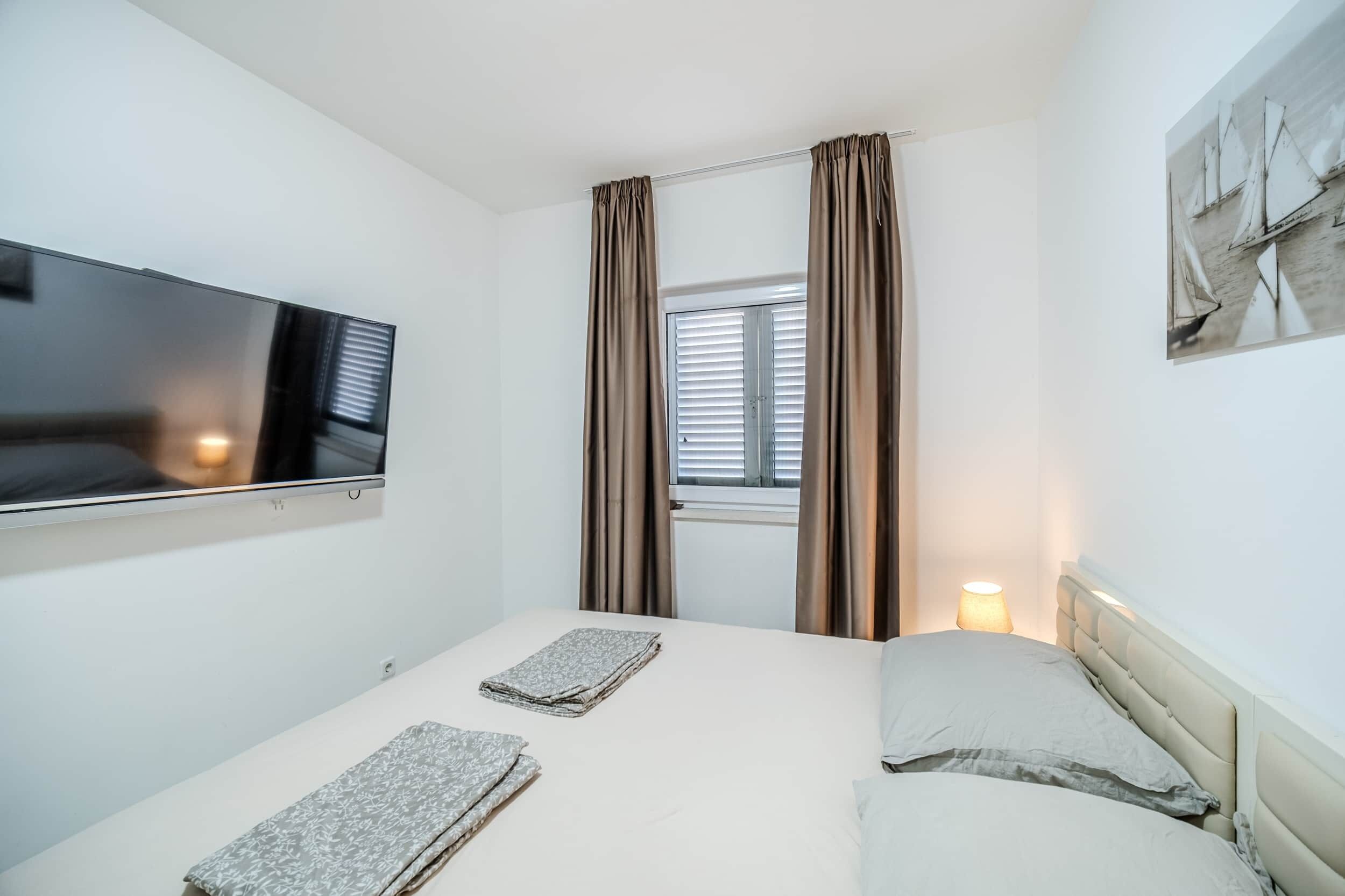 Una camera d'albergo con un letto, una televisione fissata al muro, una finestra con tende e una lampada sul comodino.