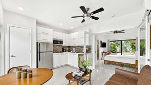 A modern kitchen with white cabinets, a microwave, and a refrigerator. There is a dining area with a wooden table and chairs, and a sofa in the living room. - Sleek Kitchenette Queen Bed (Fort Lauderdale)