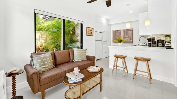 A modern living room with a brown leather sofa, a wooden coffee table, and a large window with a view of greenery. - Chic Modern Studio With Queen Bed (Fort Lauderdale)