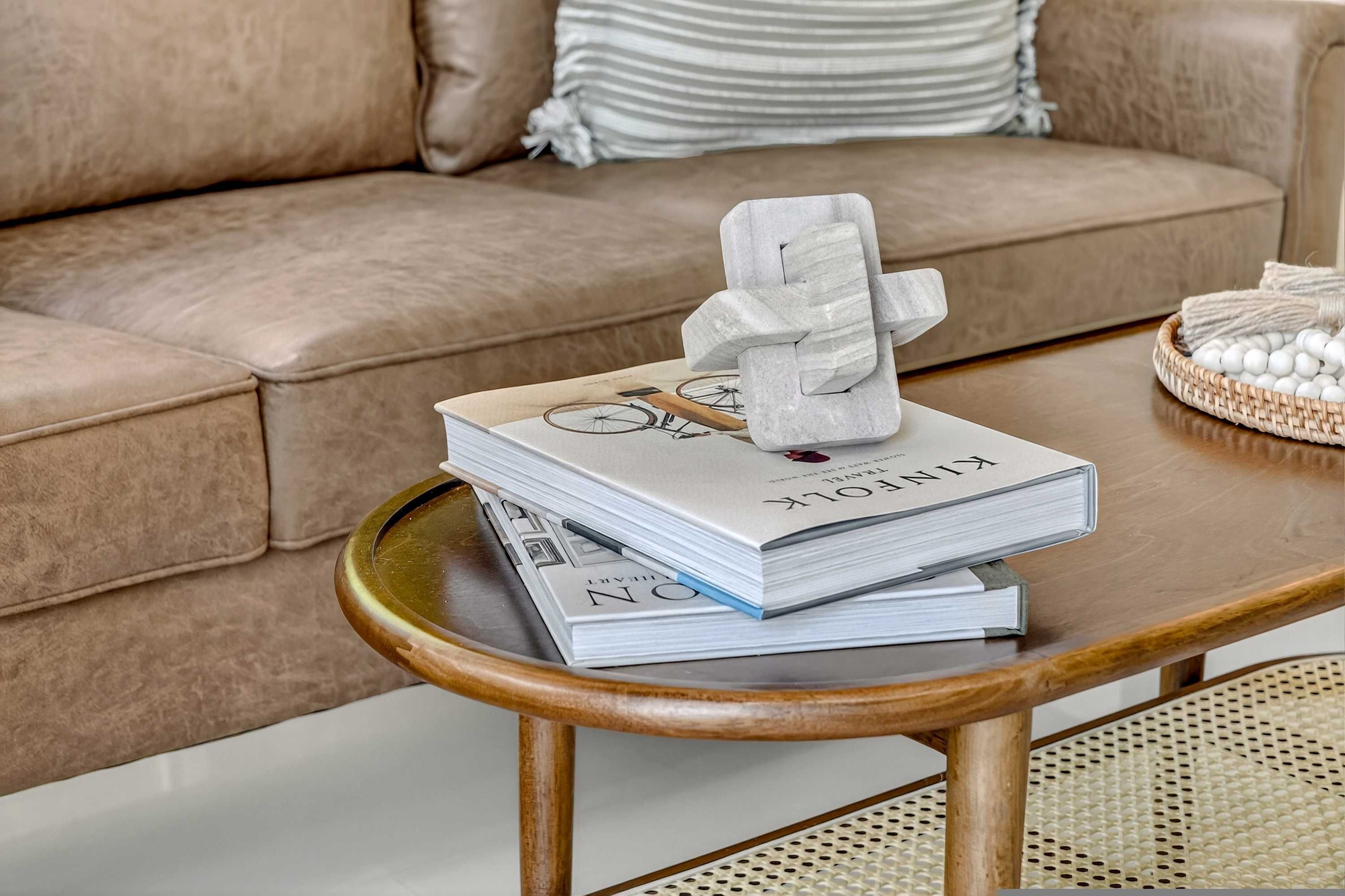 A stack of books on a wooden table with a tissue box on top, a woven basket with white balls, and a beige sofa in the background.